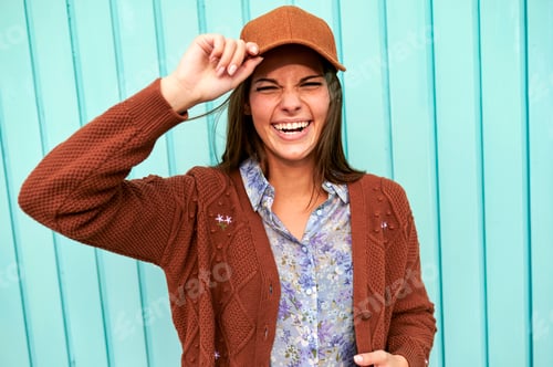 Preview: Woman laughing and holding her brown cap in front of a turquoise wall