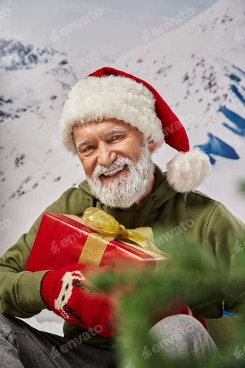 Preview: cheerful smiling Santa in christmassy hat and mittens holding present in hands, winter concept