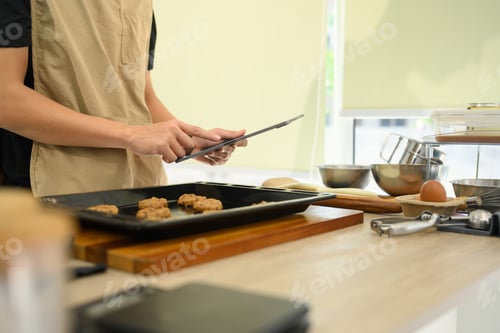 Preview: Young man preparing chocolate chip cookies in the kitchen and following recipe on digital tablet