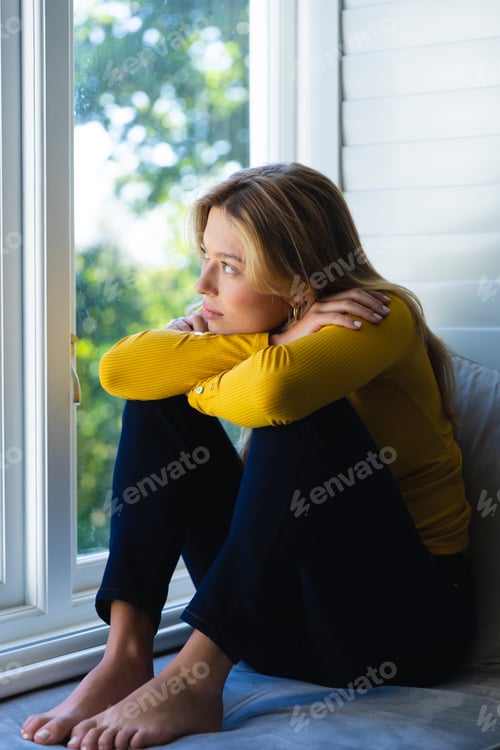 Preview: Sad caucasian woman sitting on windowsill and looking out window in sunny room at home
