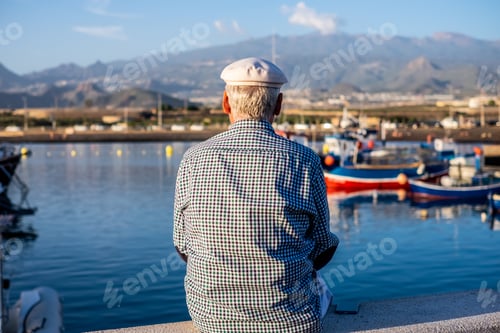 Preview: Back view of senior man in casual shirt and hat sitting at the port looking at boats and sea