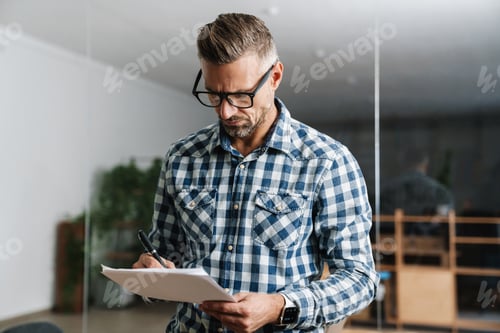 Preview: Serious white-haired man working with papers while standing in office