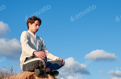 Preview: Shallow focus of a Spanish young male sitting on a rock and meditating against the cloudy blue sky