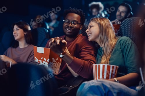 Preview: Happy couple holding hands during movie projection in a theater.