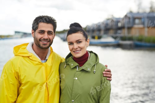 Preview: Jolly young lovers in autumn coats embracing at seaport