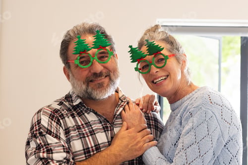 Preview: Mature couple wearing festive glasses smiling joyfully at home during Christmas