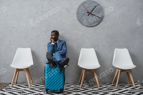 Preview: Handsome African american businessman sitting in a waiting room putting legs on suitcase