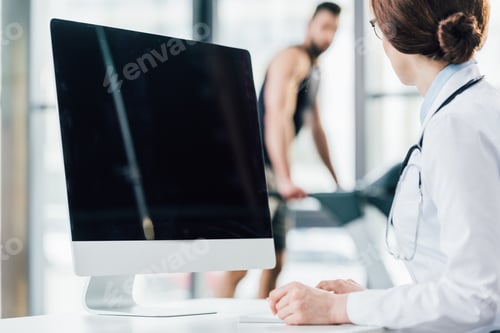 Preview: selective focus of doctor sitting near computer with blank screen in gym