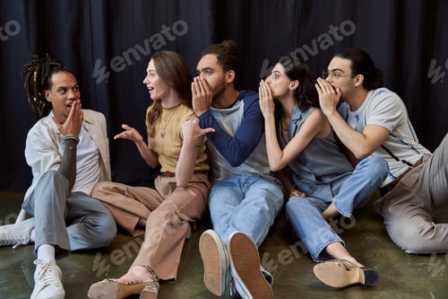 Preview: multiethnic team sitting and telling gossip to amazed colleague on black backdrop in office