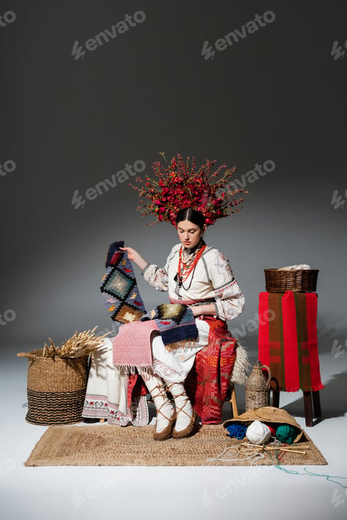 Preview: full length of brunette ukrainian woman in traditional clothes and red floral wreath holding knitted