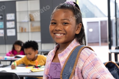 Preview: Biracial girl with a purple hair accessory smiles, wearing a backpack in a school classroom