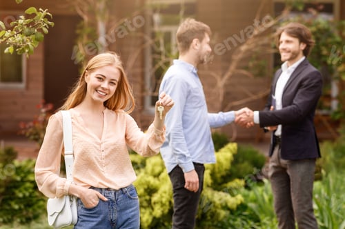 Preview: Happy young girl holding house key while her husband shaking hands with real estate agent outdoors