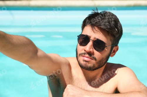 Preview: Young man wearing sunglasses taking a selfie with a smartphone in a swimming pool. Summer concept.