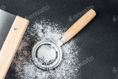 Preview: Flour scattered beside a metal sifter on a dark surface in a kitchen