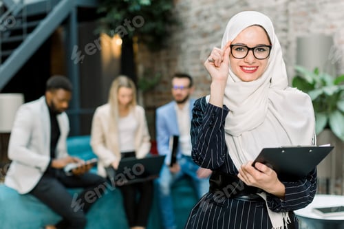 Preview: Young pretty Muslim business woman in white hijab and eyeglasses, standing in modern office, holding