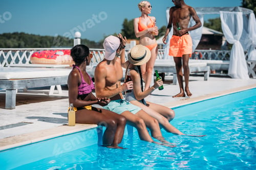 Preview: Group of young multiethnic friends sitting with beverages at poolside during vacation at resort