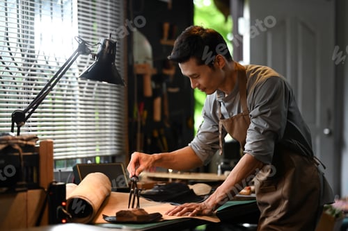 Preview: Focused Asian male shoemaker measuring piece of leather using pair of dividers on workbench