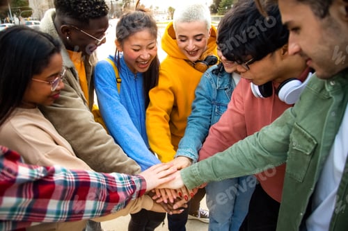 Preview: Group of multiracial college students stack hands together in a circle outdoors.