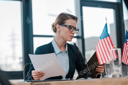 Preview: selective focus of attractive diplomat in eyeglasses looking at clipboard near american flags