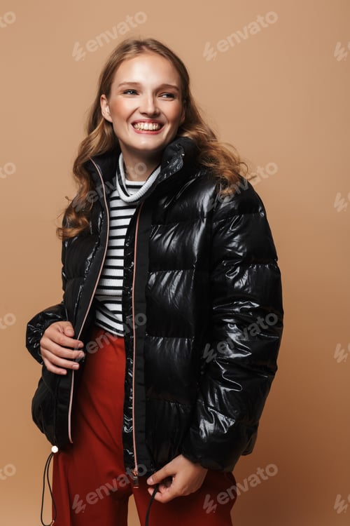 Preview: Young cheerful woman with wavy hair in black down jacket happily looking aside over beige background