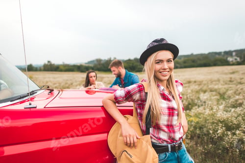 Vista previa: happy young woman leaning back on car in field during trip while her friends standing on background