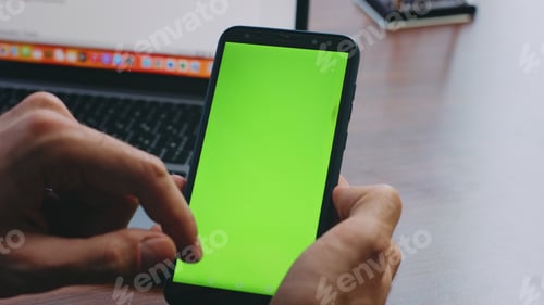 Preview: Man fingers scrolling green screen smartphone sitting desk with laptop close up.