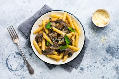 Preview: Penne rigate pasta with mushrooms, parmesan cheese and basil leaves in ceramic dish on light old con