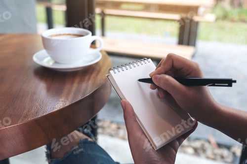 Preview: Closeup image of a woman's hands writing down on a white blank notebook with coffee cup on table