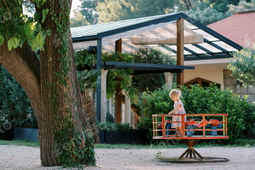 Preview: Little girl stands on a carousel in a green garden. Side view