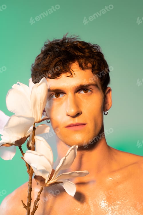Preview: Brunette man with oil on skin looking at camera near magnolia branch on green background