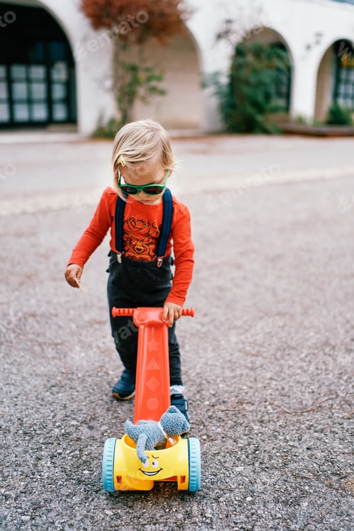 Preview: Little girl in sunglasses looks at a toy cart while standing on the road