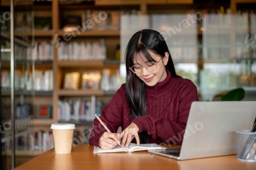 Preview: Glasses asian woman holding pencil writing in notebook aside laptop and coffee at table in library.