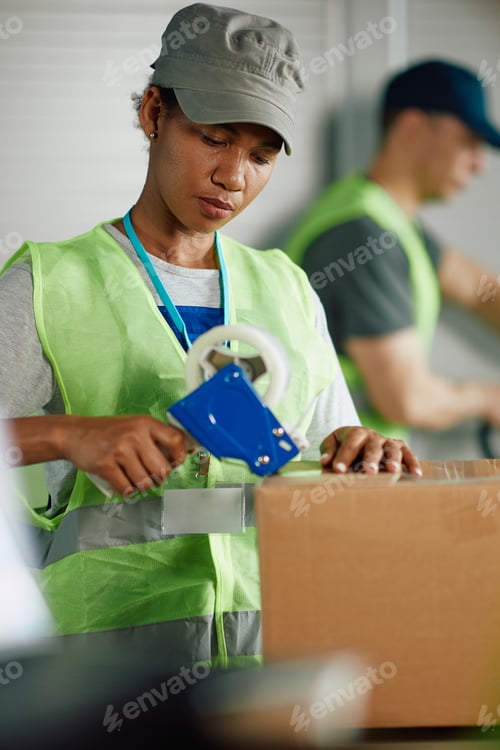 Preview: Black woman taping carboard boxes while working at distribution warehouse.