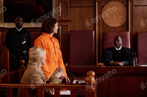 Preview: Side view of suspect in orange jumpsuit and his attorney looking at judge