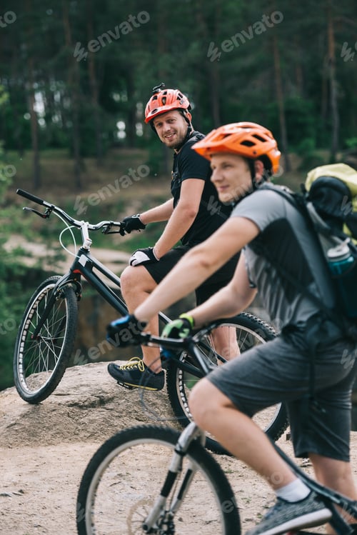 Preview: happy young trial bikers standing on rocky cliff with blurred pine forest on background