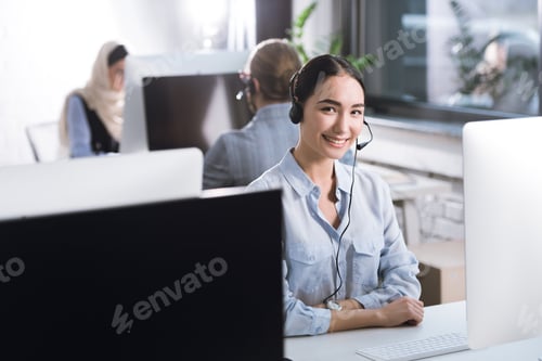 Preview: selective focus of smiling asian call center operator in headset looking at camera while sitting at