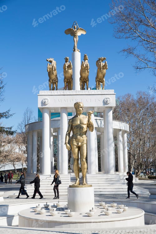 Preview: Golden statue towering in a city square as people stroll by on a sunny day