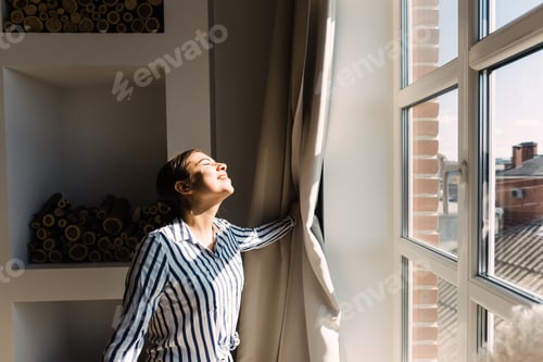 Preview: Young caucasian woman opens the curtains and looks out the window on a sunny morning
