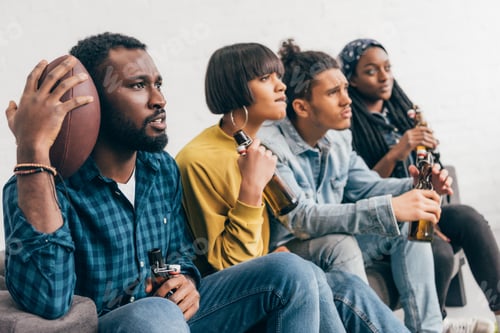 Preview: upset multicultural group of friends sitting on couch with beer bottles and watching american