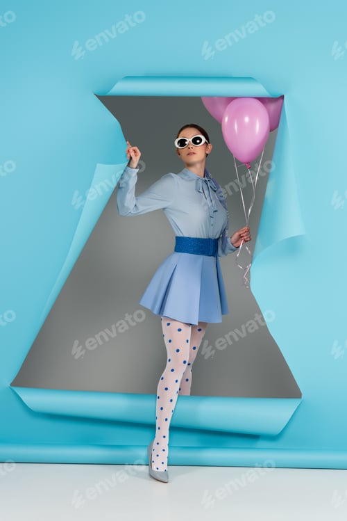Preview: extravagant woman holding pink balloons while stepping out hole in blue paper on grey background
