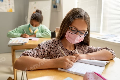Preview: Cute diligent schoolgirl in protective mask making notes in copybook