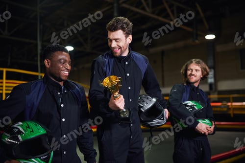 Preview: cheerful man holding golden cup and standing as a winner of go-kart race near diverse drivers