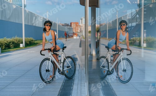 Preview: Cheerful caucasian young woman in sportswear sits on bike wearing helmet, sunglasses trains outside