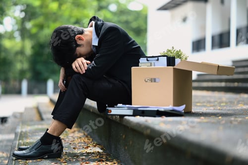 Preview: Sad, upset businessman sitting hopelessly on stairs of building with box of personal stuff.