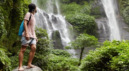 Preview: Young barefooted tourist in baseball cap standing on big stone and looking back at waterfall behind