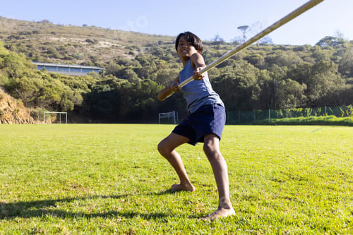Preview: Holding long pole, child boy preparing javelin throw on grassy sports field, with soccer goalposts