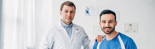 Preview: panoramic shot of smiling Physiotherapist in white coat and patient looking at camera in hospital