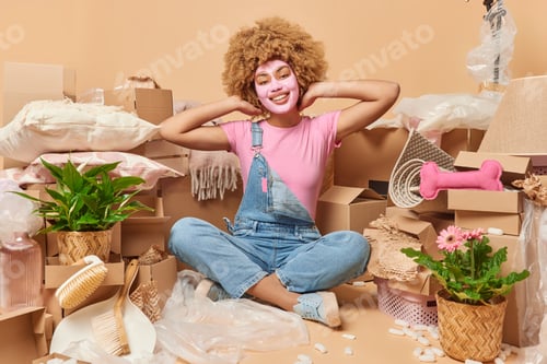 Preview: Positive good looking curly woman applies beauty mask on face sits in lotus pose dressed in t shirt