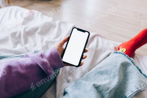 Preview: Woman holding a smartphone with a white screen mock up, resting on the bed at home.