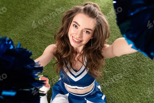 Preview: overhead view of sexy smiling cheerleader girl in blue uniform sitting with pompoms on green field